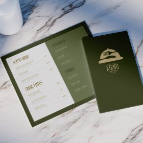 Open green restaurant menu with a matching closed cover on a marble surface.
